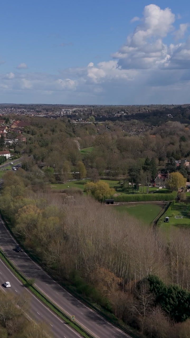 A414 roundabout vertical drone ascent over countryside roads near Hertford, England, revealing town skyline and patchwork fields under blue sky with spring clouds, light traffic. Establishing shot