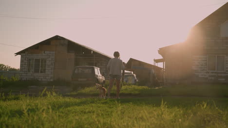 dog owner walking alongside dog on grassy field under bright sunlight, residential buildings and parked cars in background creating warm rural glow, peaceful and leisurely stroll