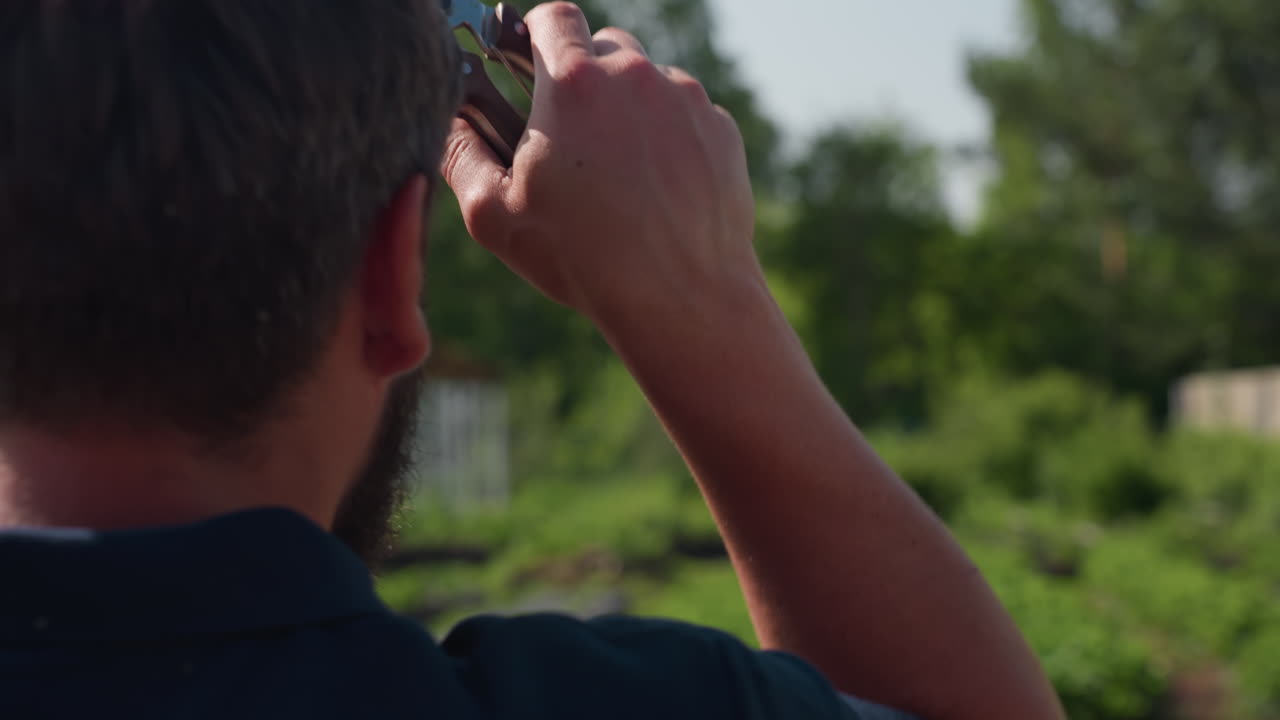 close up rear view of man on farm wiping forehead with plier in hand while inspecting green crops and white support frames under sunny sky