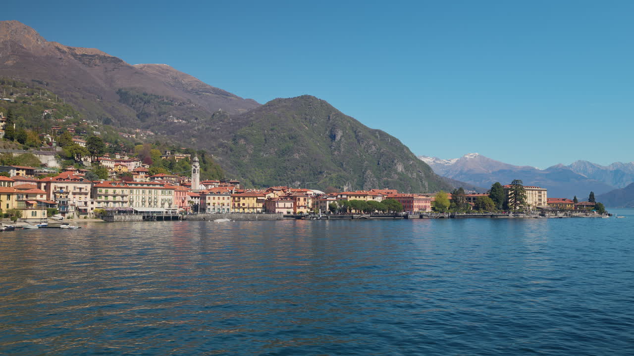 Una ripresa fluida dall'acqua rivela il colorato lungolago di una città del Lago di Como con un campanile storico, facciate color pastello, colline verdi e le Alpi innevate in lontananza in primavera