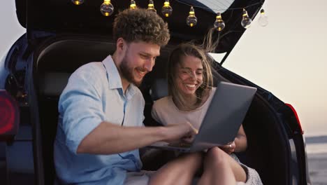 A-blonde-woman-waves-her-hand-at-a-laptop-screen-while-her-boyfriend-looks-at-a-laptop-screen-with-her-and-smiles-while-sitting