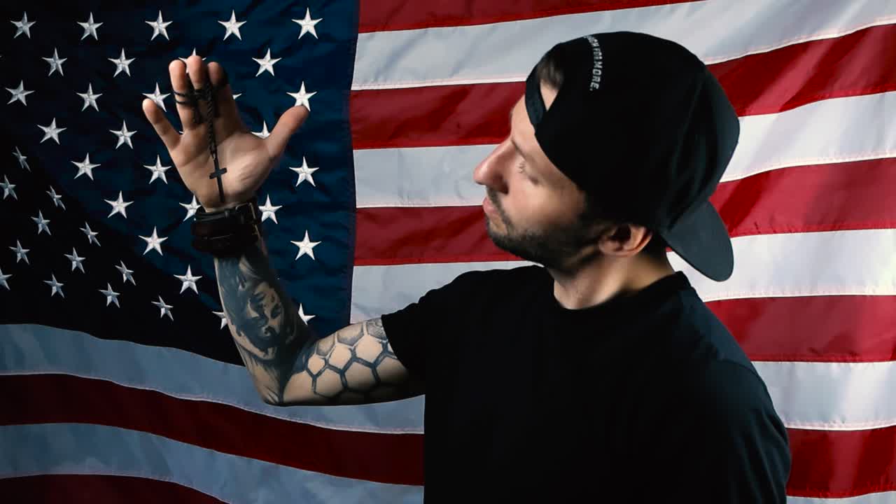 Man with full sleeve tattoo and black clothing lifting up a holy black cross in his arm on a chain, watches it and prays religiously while he is in front of an American flag. Cross moves around.
