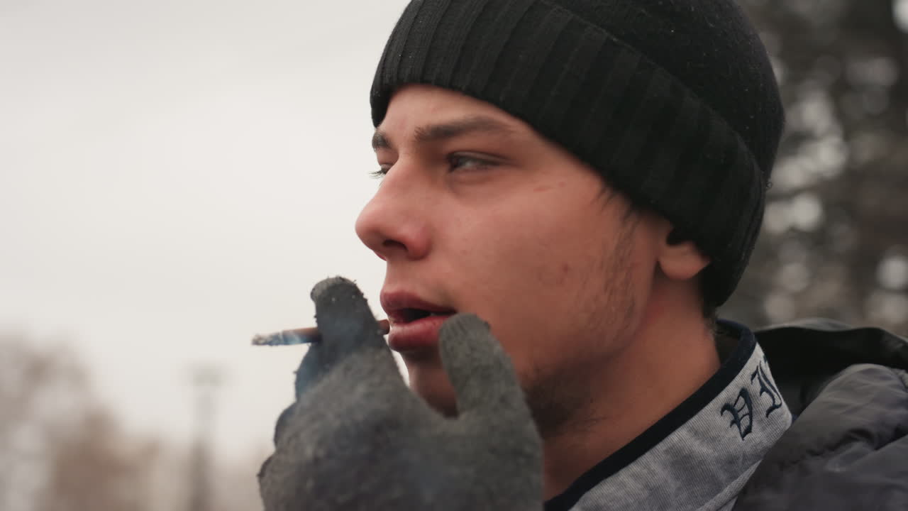 Giovane uomo bianco che fuma all'aperto in inverno, con berretto e guanti, esalando fumo denso in un parco urbano innevato, espressione pensierosa, primo piano di profilo, giacca con cappuccio, alito freddo, momento spontaneo di un pendolare.