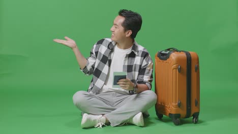 full body of asian male traveler with luggage and passport smiling and pointing to side while sitting in the green screen background studio