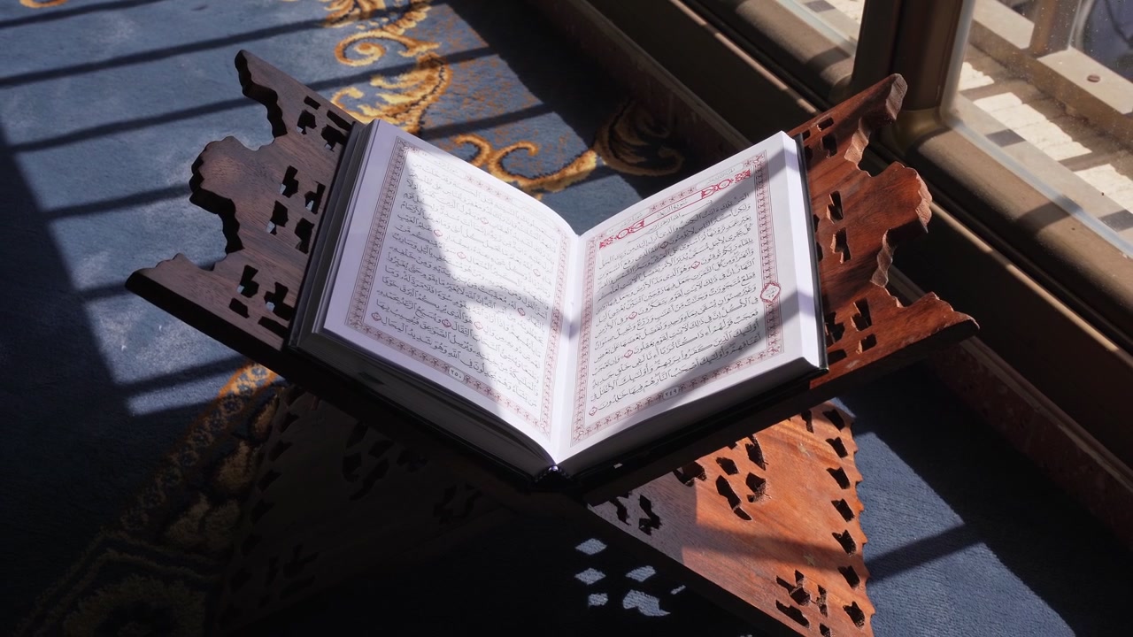 Un Sacro Corano aperto poggia su un leggio di legno intagliato all'interno. La luce del giorno intensa filtra attraverso una finestra, proiettando forti ombre sul testo arabo e sul tappeto blu a motivi della moschea.