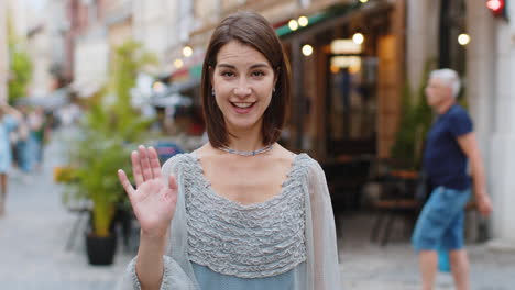 young woman smiling friendly at camera, waving hands hello, hi, greeting or goodbye in city street