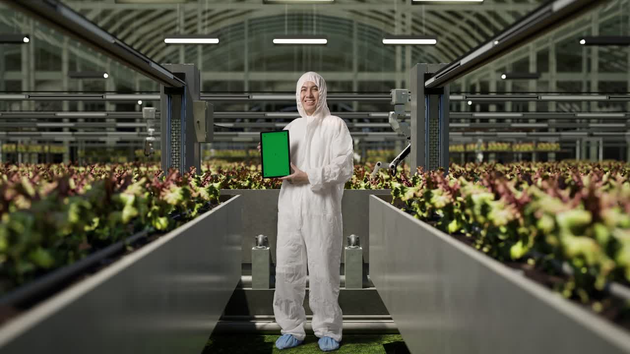 cuerpo lleno de hombre asiático investigador sonriendo y mostrando pantalla verde tableta a la cámara mientras estaba de pie en el invernadero con agricultores robóticos inteligentes