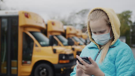 A-schoolboy-with-a-protective-mask-stands-near-a-row-of-yellow-school-buses,-uses-a-smartphone