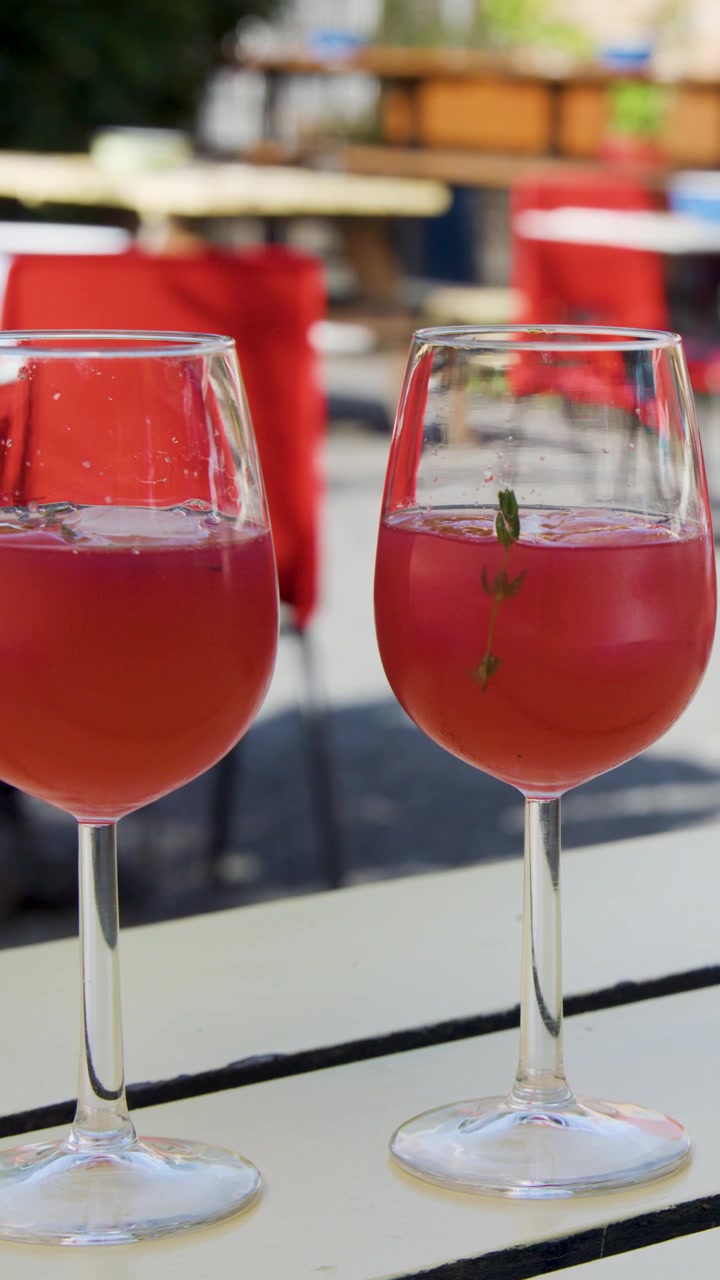 Due mani brindano con calici di vino pieni di un cocktail estivo rosso a un tavolo di un caffè all'aperto, con luce naturale e profondità di campo ridotta.