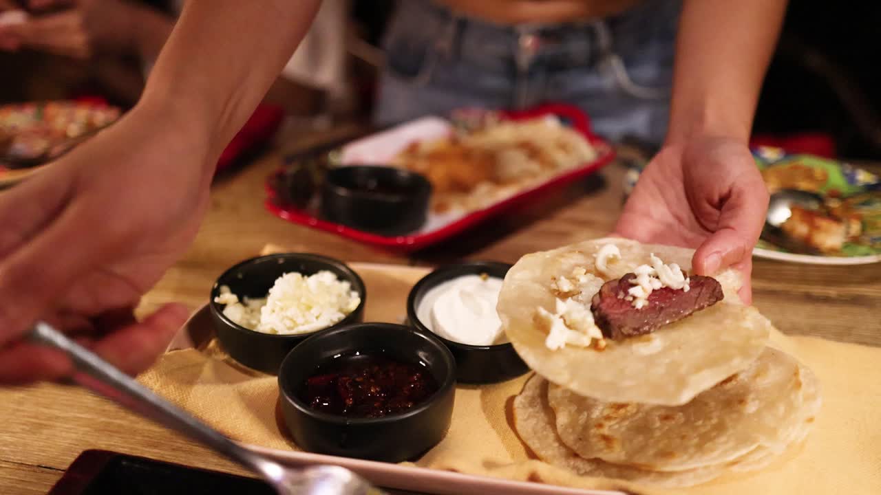 Hands assembling tlayudas with various toppings in a warmly lit restaurant setting, highlighting Mexican culinary techniques and vibrant ingredients