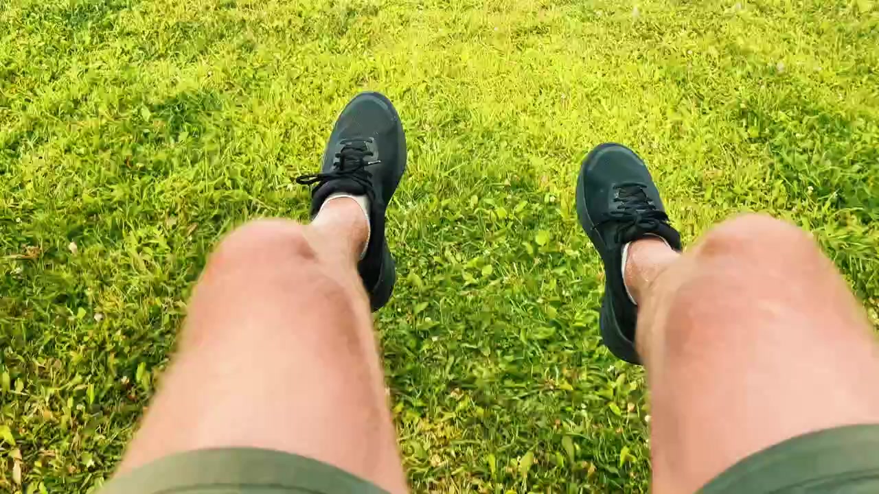 Point of view shot of person swinging legs wearing black sneakers over green grass, suitable for fitness, outdoor activity, and summer lifestyle content.
