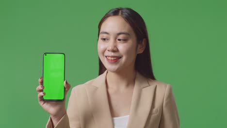 close up of asian business woman with a smile showing green screen mobile phone while standing in front of green screen