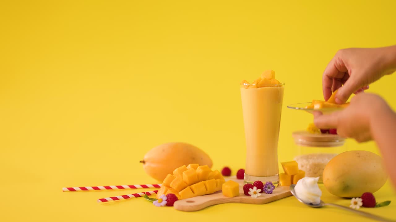 Una mano umana decora un frullato di mango alto con cubetti di frutta su uno sfondo giallo brillante in studio