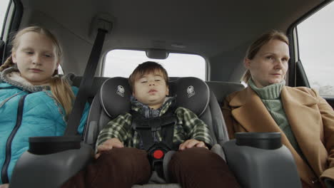 a woman with two children is driving in the back seat of a car. a kid in a child car seat and an older sister next to her