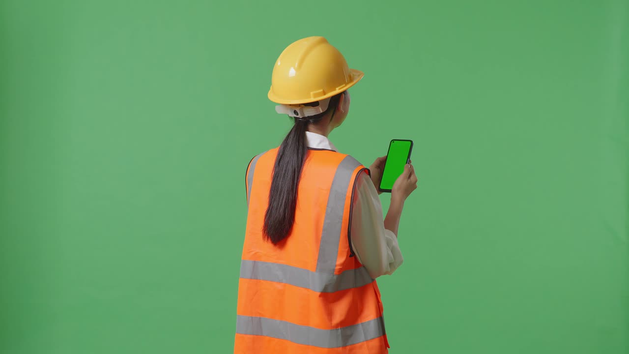 Back View Of Asian Female Engineer With Safety Helmet Working On A Green Screen Smartphone And Looking Around While Standing In The Green Screen Background Studio