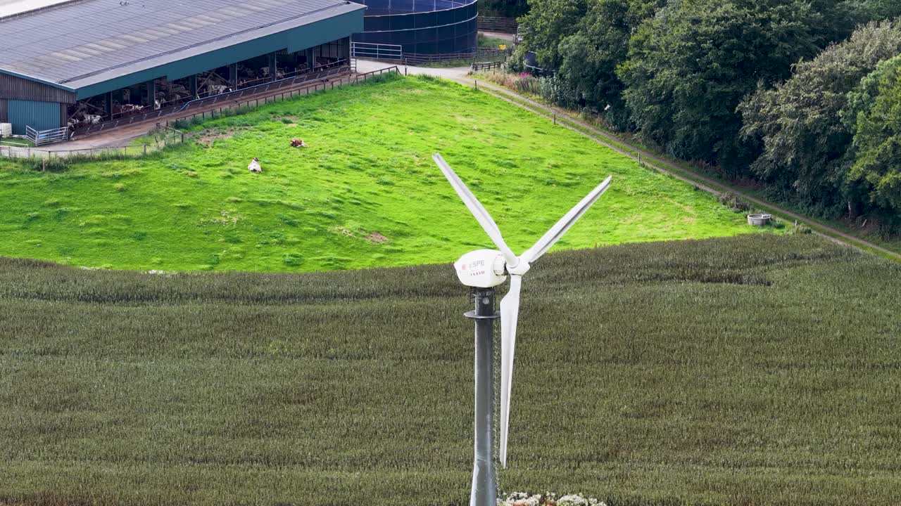 Aerial footage captures a wind turbine spinning steadily above green fields and farm buildings in Kinross, Scotland, under bright natural daylight