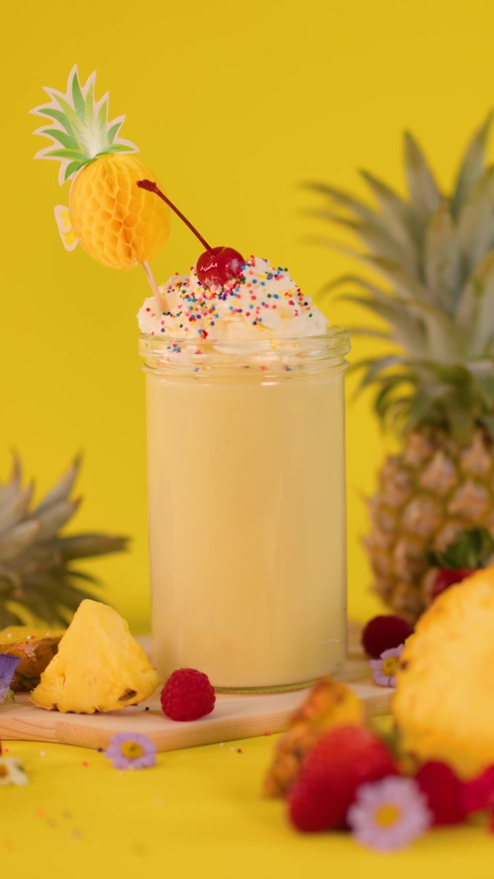 Una mano aggiunge un pezzo di ananas come guarnizione a un frullato cremoso con panna montata, codette e una ciliegia su sfondo giallo.