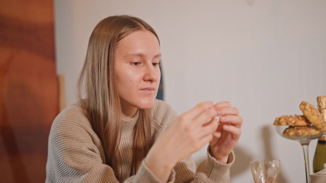 Mulher caucasiana provando croissant folhado na mesa do café da manhã, descascando camadas da massa com dedos cuidadosos, louça vintage e copo por perto, luz quente da cozinha, expressão focada, manhã doméstica