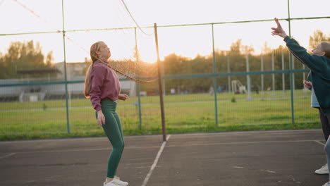 damas practicando voleibol, con una dama en leggings verdes perdiendo la pelota y persiguiéndola, el fondo presenta un campo abierto y personas involucradas en actividades durante la sesión de entrenamiento