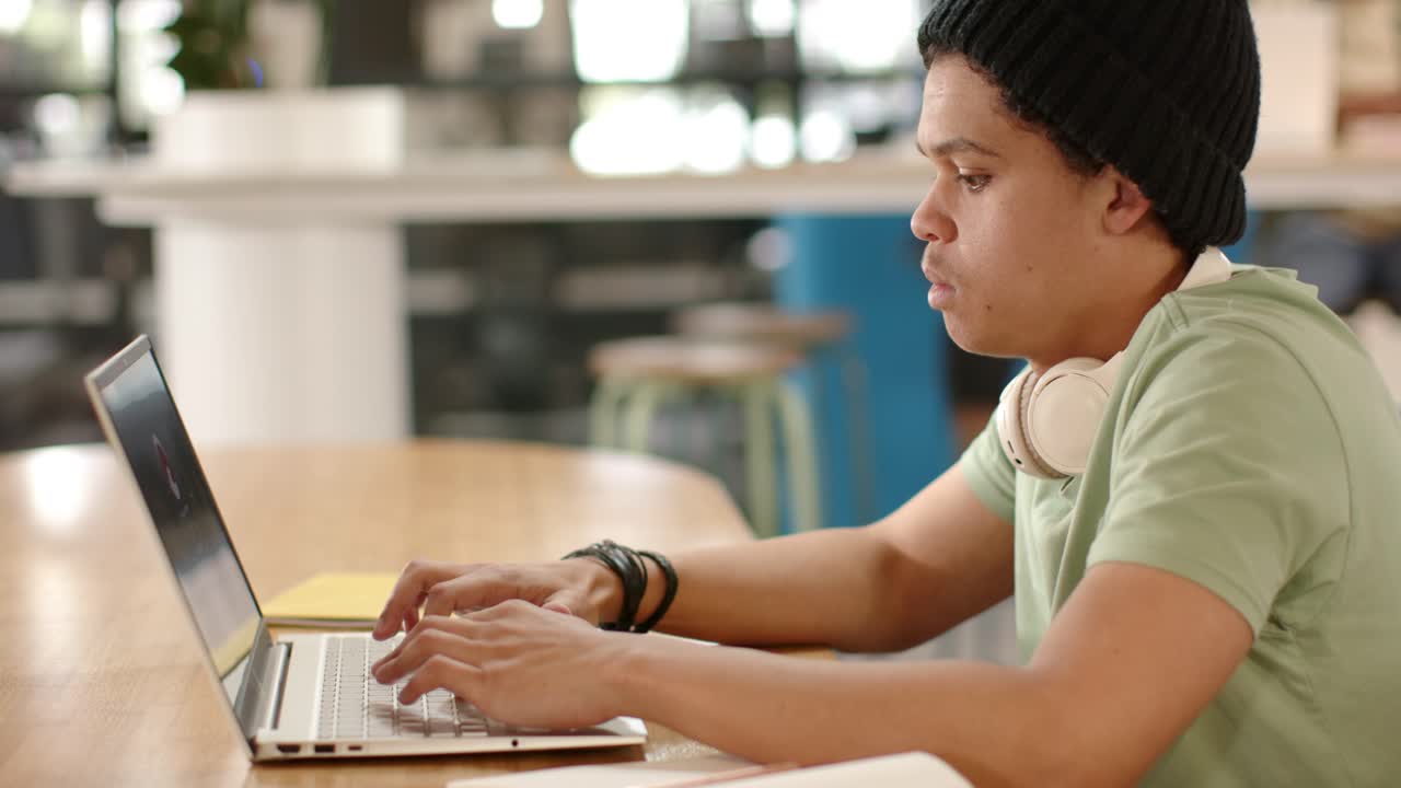 African American man typing on laptop at long table, using notebook while working with headphones