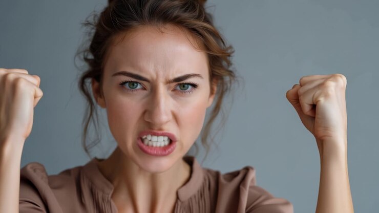 Expression intense de colère et de frustration d'une jeune femme, montrant des poings serrés et un regard déterminé, dans un décor au fond neutre.