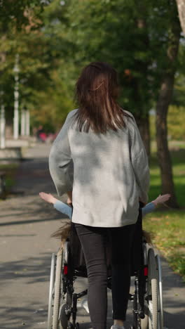 mom entertains child with disability pushing wheelchair while running on road in park. girl spreads hands to sides enjoying spring day backside view