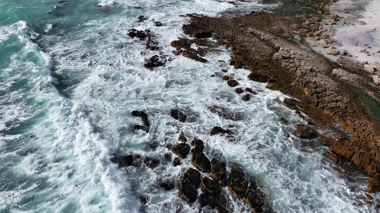 Volo di drone sopra onde turchesi che si infrangono su una spiaggia rocciosa di Città del Capo.
