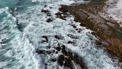 Drone-flight-above-turquoise-wave-swell-washing-in-on-rugged-Cape-Town-beach