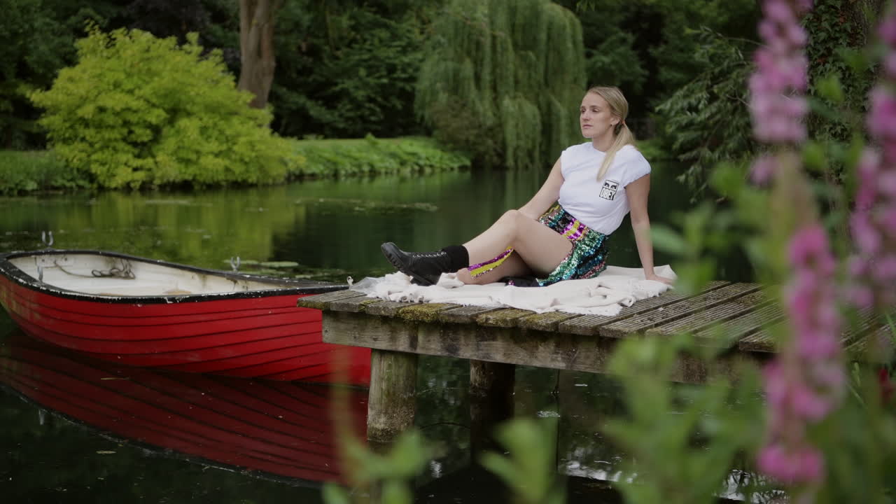 Revealing Shot Behind Flowers Of A Sexy Lady Sitting On The Jetty By The Lake
