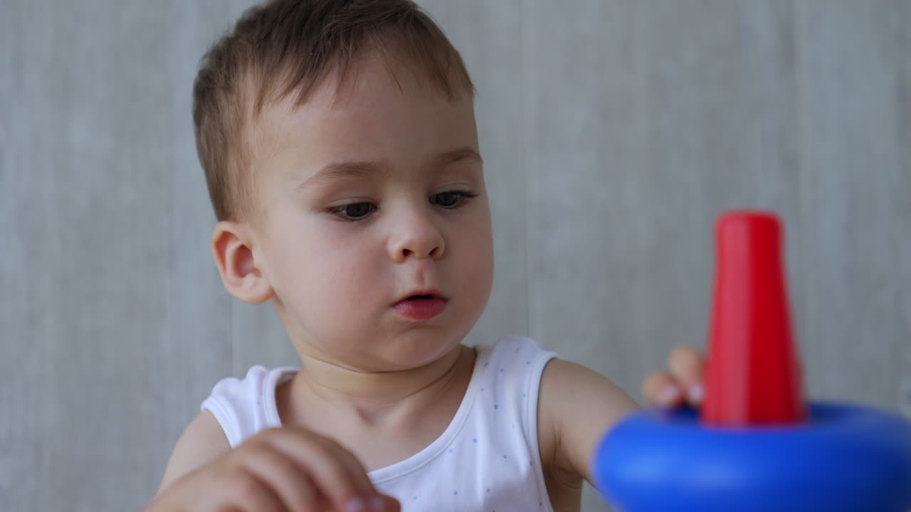 Beautiful Caucasian baby boy in white bodysuit sits on the floor. Kid plays with pyramid assembling it. Close up.