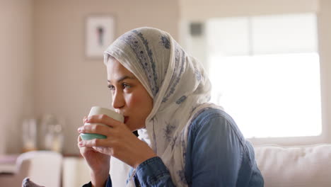 Happy-biracial-woman-in-hijab-drinking-tea-on-sofa-at-home-with-copy-space,-slow-motion