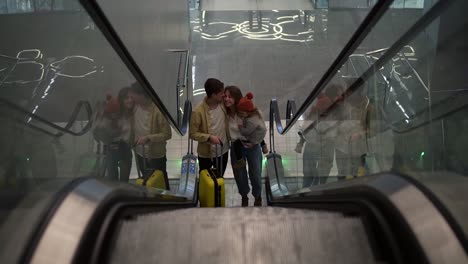 happy, young family with yellow suitcase going up by the escalator from the airport hall. smiling mother holds pretty daughter