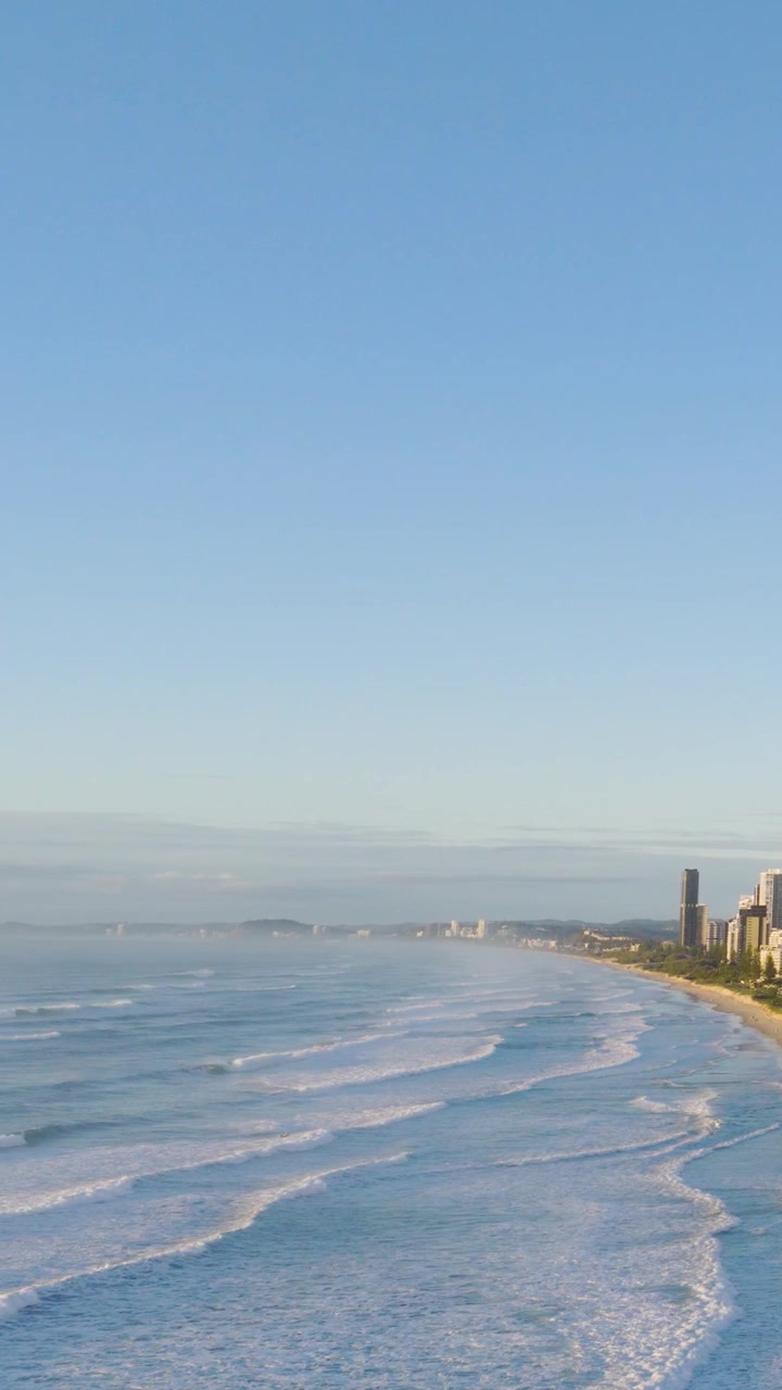Rekaman udara garis pantai Gold Coast dengan ombak, gedung pencakar langit, dan langit cerah. Menangkap suasana pantai yang tenang dan lanskap perkotaan