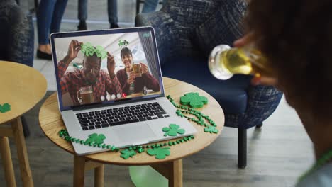 una pareja diversa sonriente con cerveza vistiendo artículos en forma de trébol en una videollamada en una computadora portátil