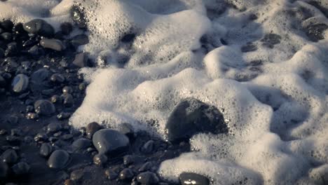 close up, slow motion view of waves from ocean washing over wet stones on beach of tenerife with sunlight shining on foaming water