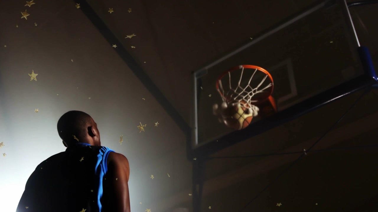 Giocatore di basket maschio che alza e rilascia la palla per segnare in palestra, con particelle a forma di stella sovrapposte al giocatore.