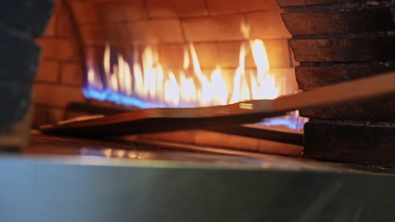Inde i en professionel stenovn danser levende blå og orange gasflammer i baggrunden, mens en pizzaspade skubber dej ind på den varme stenbund til autentisk håndværksbagning.
