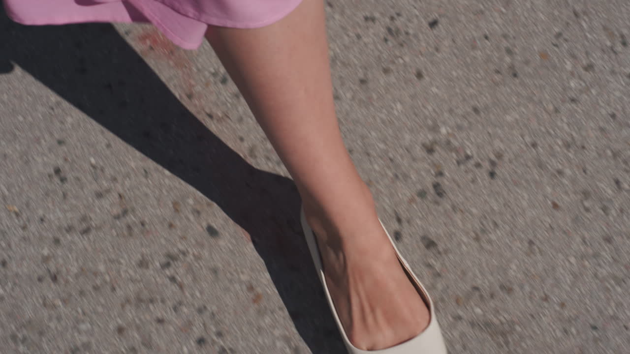 Ballerine bianche a punta su pavimentazione illuminata dal sole, orlo di un vestito rosa che sfiora l'asfalto, passi lenti e sicuri che proiettano un'ombra lunga, primo piano della caviglia e della scarpa, eleganza estiva casual con texture urbana e minimalista