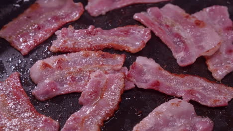 chef uses fork to flip bacon slices cooking in hot frying pan on stove