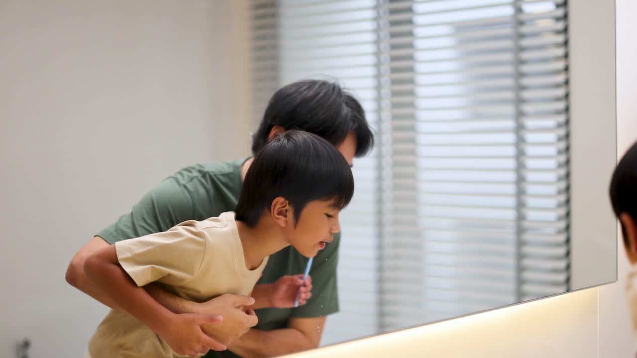 An Asian father helps his young son brush teeth at a bathroom sink, using gentle guidance and encouragement under soft, natural lighting with a steady camera
