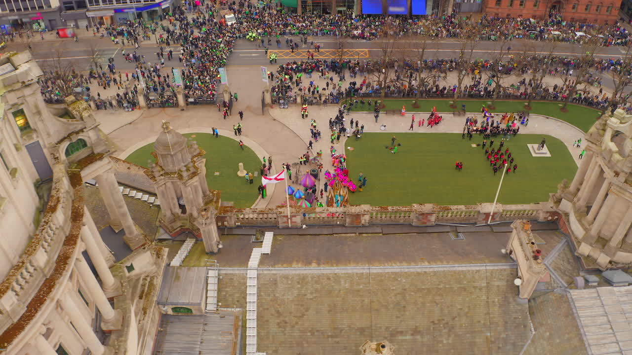 Aerial pan showcasing Belfast Town Hall's dome surrounded by gardens filled with performers, children in formation, and spectators awaiting the St. Patrick's Day parade.