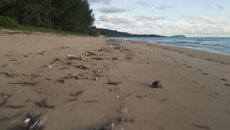 Low-aerial-backwards-flying-over-rubbish-washed-out-on-beach,-Khao-Lak,-Thailand