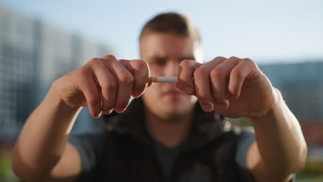 man extends hand forward holding cigarette with face and background slightly out of focus featuring modern buildings under clear daylight in urban environment