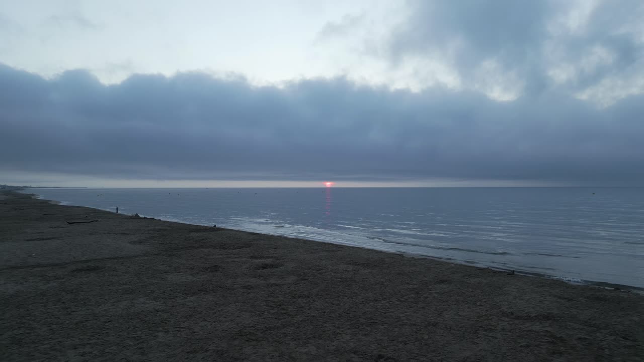 Stunning sunset casts vibrant hues over a cloudy beach. Waves crash gently on the shore while a few people stroll along the sandy coastline, enjoying the serene evening atmosphere