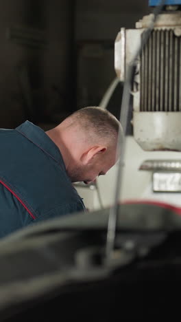 ingeniero en uniforme azul trabajando en un taller mecánico con un coche blanco estacionado en el fondo, entorno industrial con herramientas y equipos, centrado en la tarea de reparación en un espacio de trabajo organizado