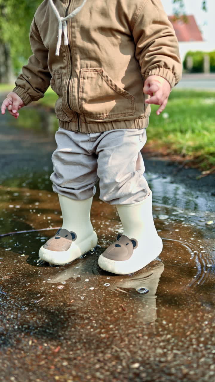 Boy in a panama hat and beige jacket on a walk after the rain. Cute 2 year old boy in rubber boots runs through puddles after the rain. Child jumps in a puddle on the street