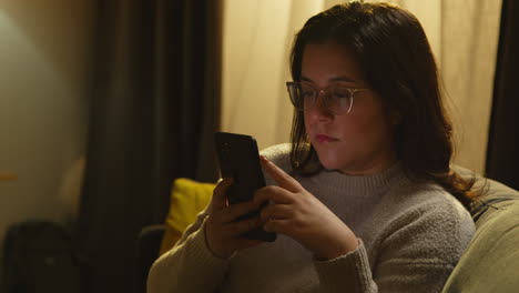 young woman at home sitting on sofa with mobile phone scrolling through internet or social media in the evening 2