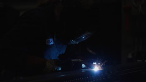 slow mo, blue collar worker welding metal, bright sparks in dark workshop room