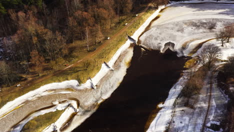 Gauja-river-in-Valmiera-is-frozen-during-winter-time,-snowy-riverbank-and-forest