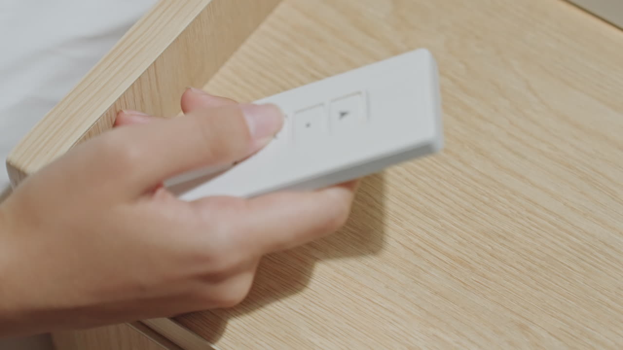 Guest reaches for bedside remote, delicate ring on finger, white sleek control placed on wooden nightstand, soft linens visible, gentle morning light, finger presses button to adjust lighting, quiet